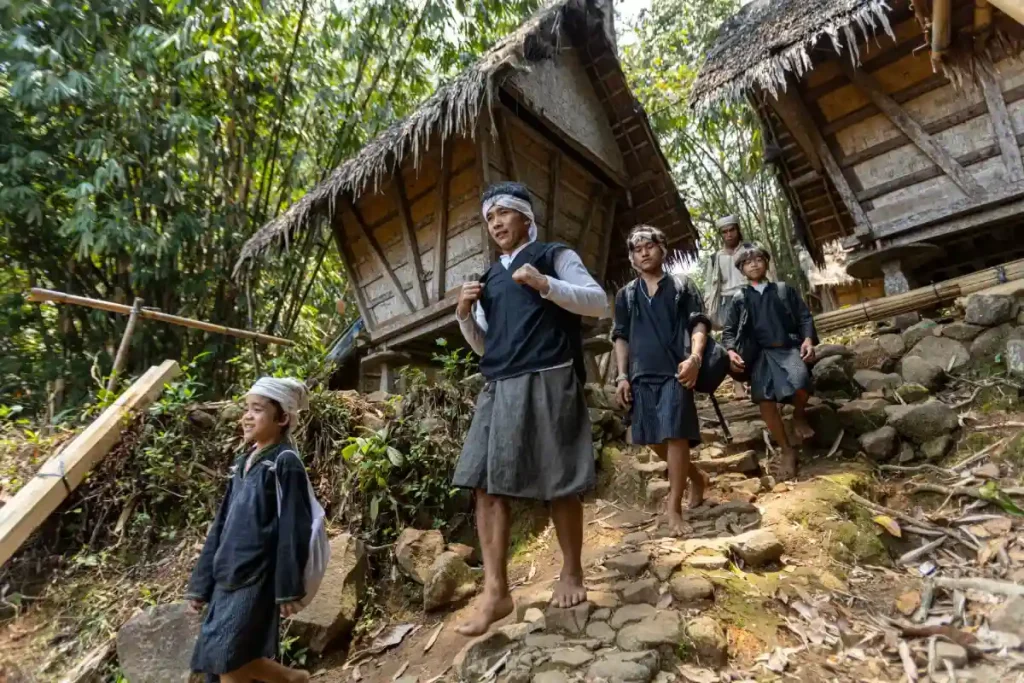 Sekelompok warga Suku Baduy Dalam berjalan di jalan berbatu di antara rumah-rumah tradisional berdinding bambu dan beratap ijuk, mengenakan pakaian adat berwarna hitam dan putih di lingkungan pedesaan yang dikelilingi pepohonan hijau.